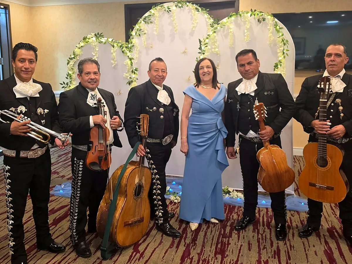 El mariachi acompañando a una señora de vestido azul ante un arco floral en un salón